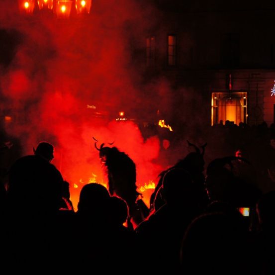 Eine Nachtszene mit einer Menschenmenge, die sich um ein Feuer versammelt, das rote Rauchwolken ausstößt, mit Menschen in Hörnerkostümen.