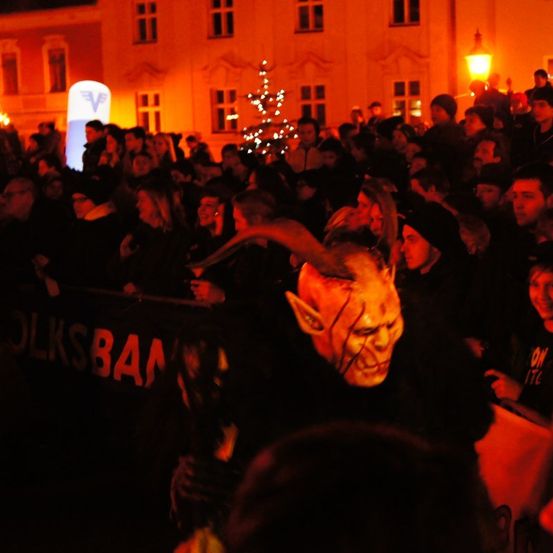 Eine große Menschenmenge versammelt sich bei Nacht im Freien, einige tragen Masken, beleuchtet von roten Lichtern, mit einem Weihnachtsbaum im Hintergrund.