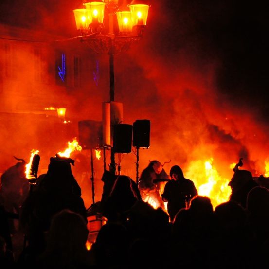 Eine Menschenmenge beobachtet eine feurige Aufführung vor einem Gebäude bei Nacht. Zwei Personen spielen Musikinstrumente in der Nähe des Feuers.