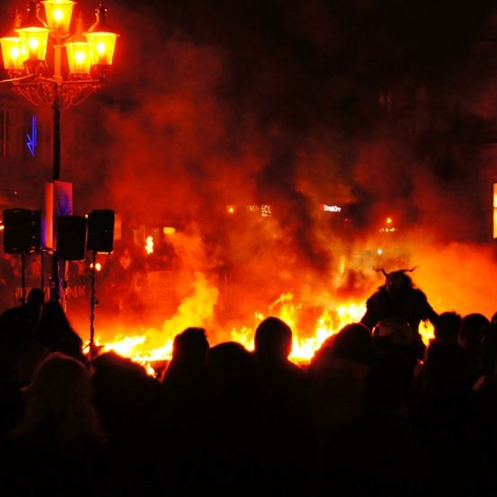 Ein Lagerfeuer brennt in der Nacht hell, während eine Menschenmenge zuschaut. Eine Person mit einer Hörnermaske steht in der Nähe des Feuers.