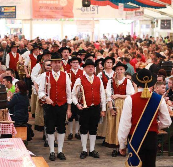 Eine Gruppe von Musikern und Teilnehmern befindet sich auf einem Festival, wo sie auftreten und Essen und Getränke genießen. Die Musiker tragen rote Westen und Hüte, während andere an Tischen sitzen.