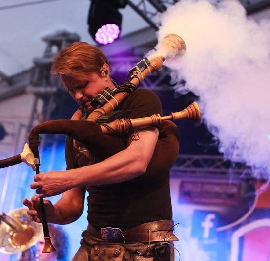 Ein Mann spielt Dudelsack auf der Bühne mit einer Trompete im Hintergrund. Er stößt Rauch aus den Dudelsack aus, während er auftritt.