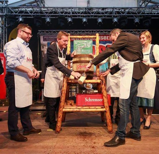 Vier Personen in Schürzen stehen um einen hölzernen Fass. Zwei drücken etwas, einer hält einen Behälter und der andere lächelt. Ein Mikrofon steht in der Nähe.