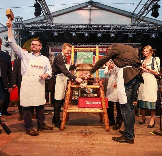 Menschen sind um eine Bühne versammelt, ein Mann hält einen Hammer, ein anderer schenkt Bier ein. Eine rote Box mit Schrenser darauf steht auf der Bühne.