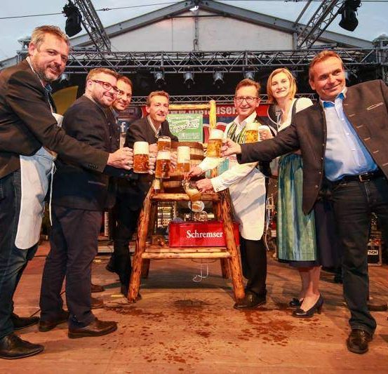 Mehrere Männer und Frauen stehen auf der Bühne, halten Biergläser und schenken Bier aus einer roten Kiste ein. Ein Mikrofon steht vor ihnen.