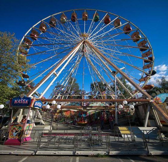 Ein großes Riesenrad mit dem Wort Piege darauf. Mehrere Fahrgeschäfte umgeben es, und eine Person befindet sich am Tickethäuschen. Bäume und ein klarer blauer Himmel im Hintergrund.