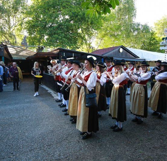 Eine Band tritt in traditioneller Kleidung auf, steht auf einem gepflasterten Bereich, mit Zuschauern und Händlern im Hintergrund.