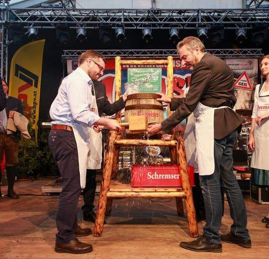 Zwei Männer in Schürzen klopfen mit einem Vorschlaghammer auf einen Fass bei einer Veranstaltung im Freien, eine Frau in einem Kleid beobachtet. Ein Mann in einem Anzug spricht in ein Mikrofon auf der linken Seite.