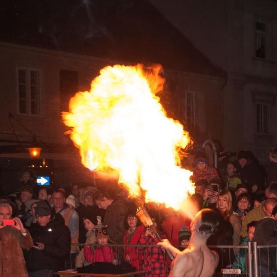 Ein Mann, der vor einer Menge Feuer bläst und dabei eine große Flamme erzeugt, während einer nächtlichen Veranstaltung. Im Hintergrund sind Gebäude und Straßenlaternen zu sehen.