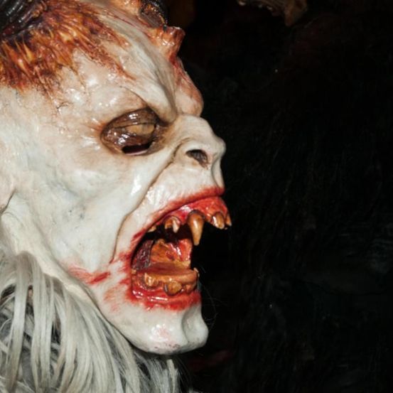 Ein Nahaufnahme einer weißen, blutigen Maske mit offenem Mund, Fangzähnen und Blut um den Mund, beleuchtet von einem dunklen Hintergrund.