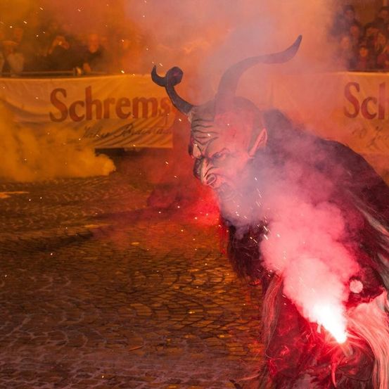 Eine Person in einem Teufelskostüm mit Hörnern bläst Feuer aus einer Fackel, während Rauch um sie herum aufsteigt. Eine Menge beobachtet hinter einem Banner mit dem Wort 'Schrems'.