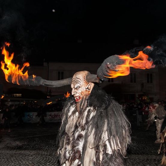 Eine Person in einem gehörnten Kostüm hält brennende Fackeln. Die Szene spielt bei Nacht mit Menschen im Hintergrund.