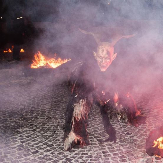 Eine Person in Teufelskostüm steht vor einem Feuer mit Rauch auf einer Kopfsteinpflasterstraße.
