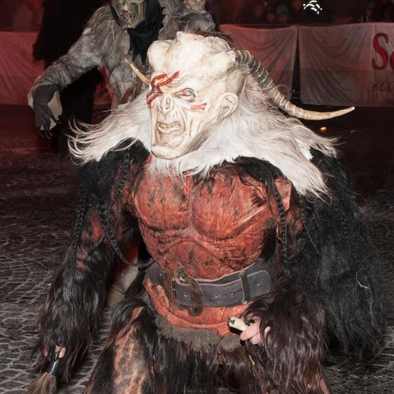 Eine Person in einem Hörnerkostüm steht auf einem Kopfsteinpflasterboden, mit einer anderen Person in einer Maske hinter ihr. Der Hintergrund ist verwischt und der Ort scheint nachts zu sein.