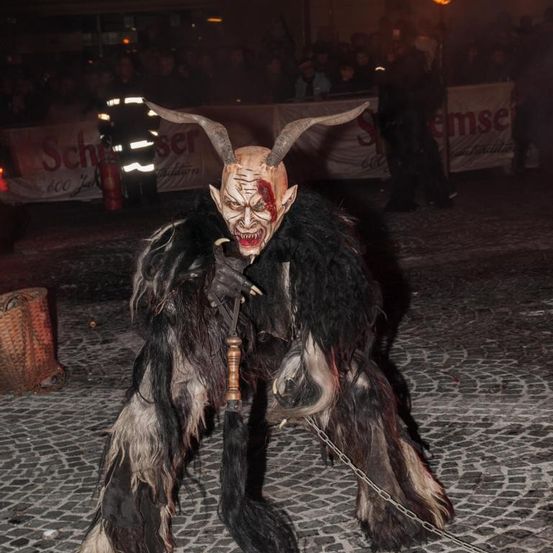 Eine Person in einem gehörnten Kostüm steht angekettet und hält eine Lanze in der Hand, vor einer Menge, mit Bannern und einem Feuer im Hintergrund.