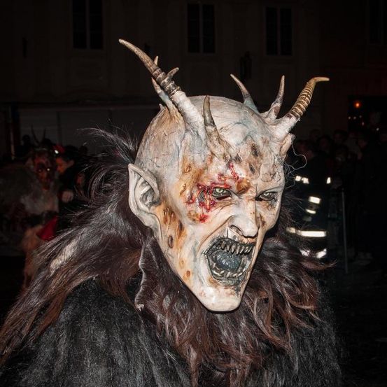 Eine Person in einem hörnertragenden Teufelskostüm steht bei Nacht in einer Menschenmenge, mit einem Gebäude und einem Feuerwehrmann im Hintergrund.