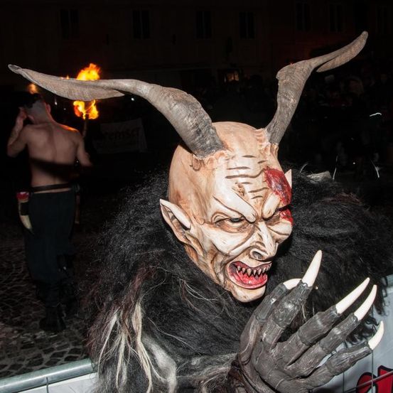 Eine Person in einem hörnertragenden Dämonenkostüm steht bei Nacht auf der Straße mit einer Menschenmenge hinter sich. Im Hintergrund hält ein Mann eine brennende Fackel.