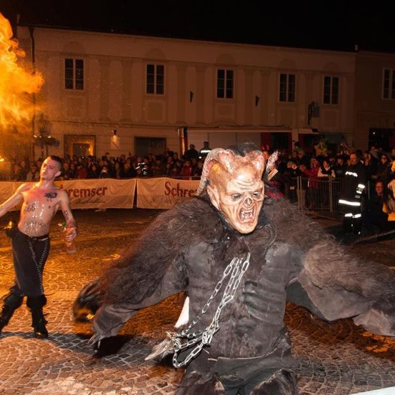 Ein maskierter Darsteller in einem Kostüm läuft an einem tätowierten Mann mit Feuer auf der Straße vorbei, vor einer Menschenmenge und einem Gebäude im Hintergrund.
