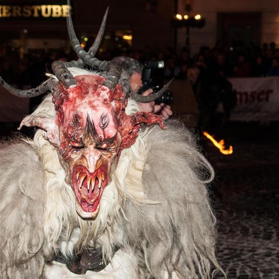 Eine Person in einem Dämonenkostüm mit Hörnern steht vor einer Menschenmenge bei Nacht. Ein Mann dahinter hält die Szene mit einer Kamera fest.