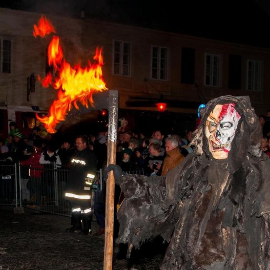 Eine Menge beobachtet, wie eine Person in einem gruseligen Kostüm mit einer Fackel vor einem Feuer steht.