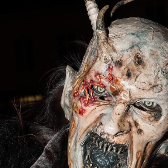 Ein Nahaufnahme eines Dämonenmaske mit grünen Augen, blutigem Gesicht und einem Horn auf dem Kopf. Die Maske wird von jemandem getragen, vielleicht für Halloween.