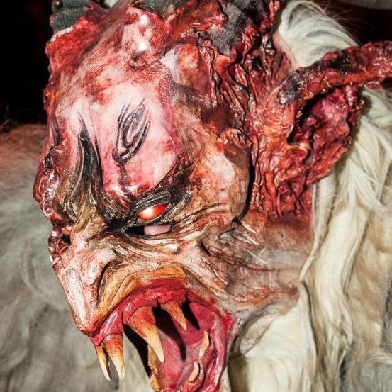 Eine Nahaufnahme eines furchterregenden, blutbefleckten Maske mit Hörnern, die ein schreckliches Gesicht mit roten Augen, scharfen Zähnen und einem bösen Ausdruck zeigt.