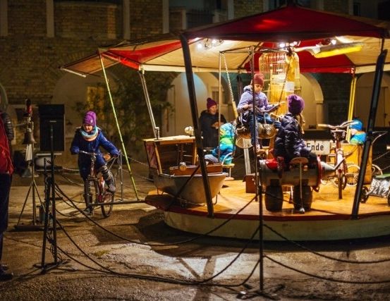 Ein Karussell mit rotem Dach ist in einem Außenbereich bei Nacht aufgebaut, einige Kinder fahren auf Fahrrädern und andere stehen herum.