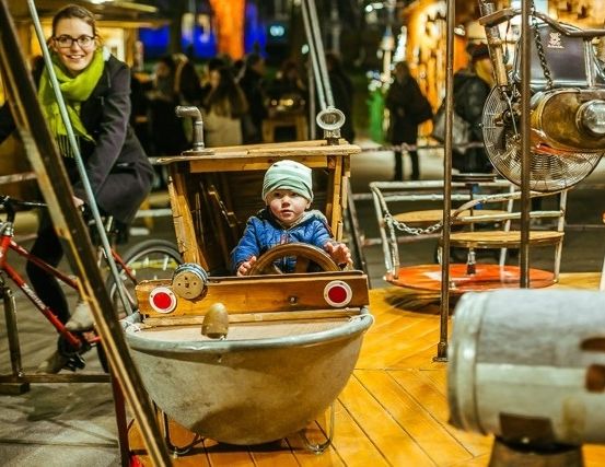 Eine Frau fährt mit einem kleinen Kind, das ein bootförmiges Fahrgeschäft steuert, ein Fahrrad. Verschiedene Metallgegenstände und Stühle sind um sie herum, mit Menschen im Hintergrund.
