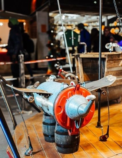 Ein Flugzeugmodell wird auf einer hölzernen Plattform mit einem roten Fass als Rumpf ausgestellt. Es steht ein Golfschläger an der Seite. Hinter dem Modell stehen Leute. Es gibt einen Metallpfahl und eine Kette, die am Modell befestigt sind. Es gibt einen verschwommenen Blick auf ein hölzerne Fass und eine Person, die dahinter steht.