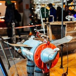 Ein Flugzeugmodell wird auf einer hölzernen Plattform mit einem roten Fass als Rumpf ausgestellt. Es steht ein Golfschläger an der Seite. Hinter dem Modell stehen Leute. Es gibt einen Metallpfahl und eine Kette, die am Modell befestigt sind. Es gibt einen verschwommenen Blick auf ein hölzerne Fass und eine Person, die dahinter steht.