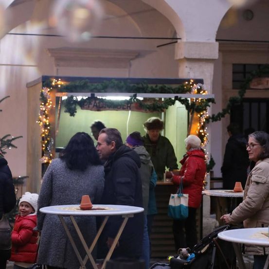 Eine Gruppe von Menschen steht um einen mit Weihnachtslichtern geschmückten Imbissstand herum. Eine Frau in einem roten Mantel steht hinter der Theke, während andere zuschauen. Eine Frau mit einem Kinderwagen steht in der Nähe.
