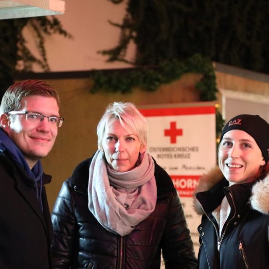 Drei Personen stehen vor einer Wand mit einem Schild mit einem roten Kreuz und Text und lächeln in die Kamera.