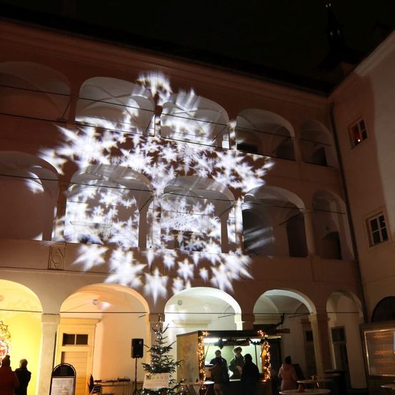 Bei Nacht projiziert ein Gebäude mit Bogenfenstern ein Schneeflockendesign auf seine Fassade. Ein beleuchteter Weihnachtsbaum steht davor, und Menschen sind in der Nähe versammelt.