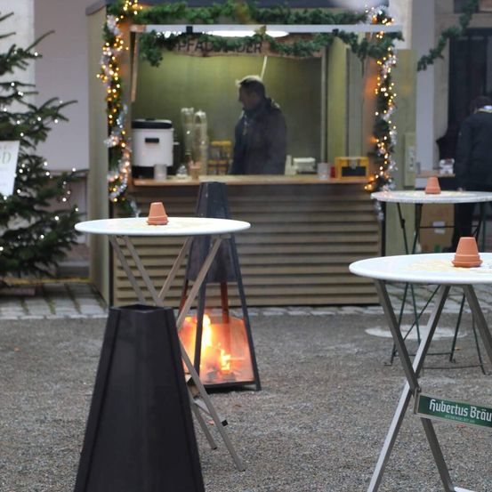 Ein Weihnachtsmarkt-Aufbau mit einem kleinen Tisch und einem brennenden Feuerkorb davor. Eine Person steht hinter dem Tresen im Inneren, mit ausgestellten Artikeln.