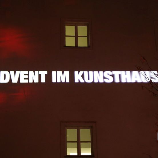 Ein Gebäude mit dunkler Außenseite hat eine Projektion von weißem Text, der 'ADVENT IM KUNSTHAUS' lautet. Es gibt zwei Fenster mit warmem Licht im Inneren.