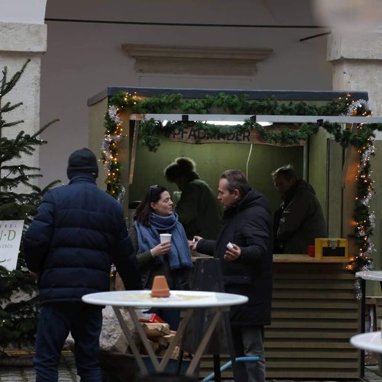 Ein Mann und eine Frau unterhalten sich vor einem geschmückten Stand. Die Frau hält eine Tasse, und der Mann einen Spielcontroller. Sie stehen vor einem Tisch mit einem Topf.