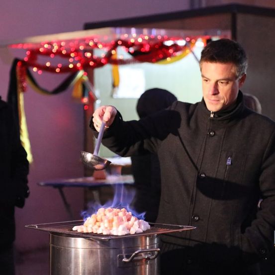 Ein Mann in einer Kochjacke kocht Marshmallows über einem Feuer auf einem Grill, mit dekorativen Lichtern im Hintergrund.