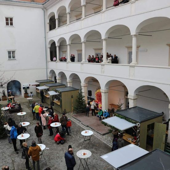 Eine Vogelperspektive eines Innenhofs mit Menschen, die um Tische, einen Weihnachtsbaum und Essensstände versammelt sind. Der Innenhof ist von gewölbten Gängen und Balkonen mit stehenden Personen umgeben.