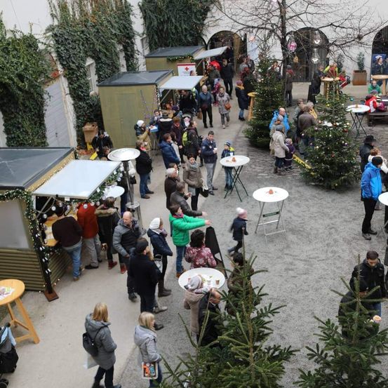 Eine Luftaufnahme eines Innenhofs während der Wintersaison mit Menschen, die herumlaufen und an Tischen sitzen, umgeben von Weihnachtsbäumen und Ständen.