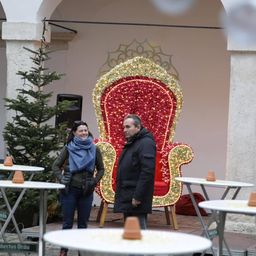Zwei Personen stehen draußen, eine Frau in einem blauen Schal und ein Mann in einem schwarzen Mantel, vor einem großen dekorativen Stuhl mit rotem und goldenem Glitzer. Ein Weihnachtsbaum steht hinter ihnen, und es sind Tische und Stühle im Bereich aufgestellt.