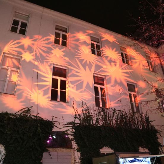 Ein weißes Gebäude mit mehreren Fenstern ist bei Nacht mit einer orangefarbenen sternförmigen Lichtinstallation an der Wand beleuchtet.