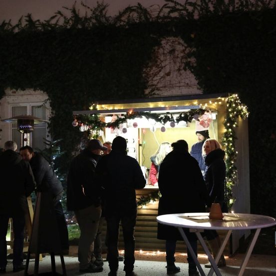 Menschen versammeln sich bei Nacht um einen festlich geschmückten Kiosk, der mit Weihnachtslichtern und Grün behängt ist, während andere stehend von der Seite beobachten.