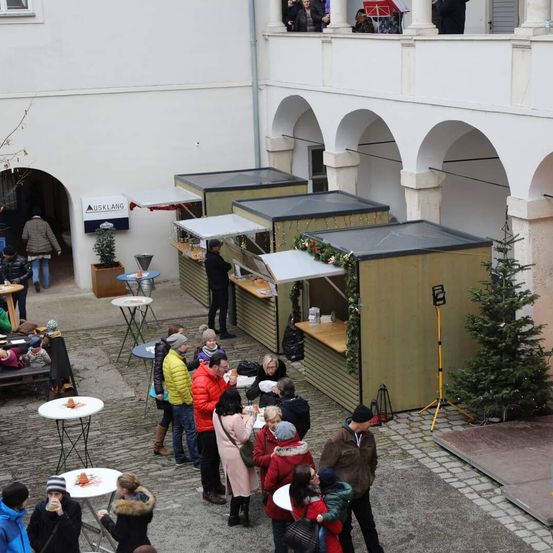 Eine Gruppe von Menschen steht in einem Außenhof mit kleinen Essensständen und einem Weihnachtsbaum. Die Stände haben Markisen und Dekorationen, und einige Menschen essen an den Tischen. Oben hat ein Balkon Topfpflanzen und Menschen.