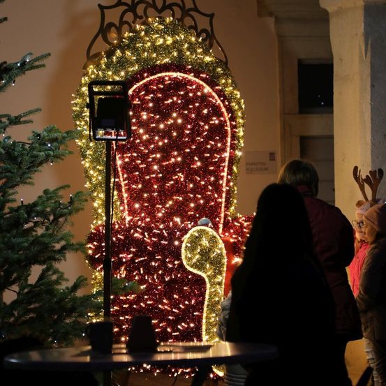 Ein geschmückter Schlitten mit roten und gelben Lichtern steht prominent im Vordergrund neben einem Weihnachtsbaum. Im Hintergrund versammeln sich Menschen in der Nähe eines Gebäudes mit offener Tür.