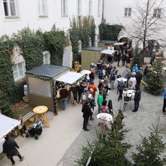 Eine Vogelperspektive auf einen Innenhof mit mehreren Menschen, die gehen und sitzen. Einige tragen Winterkleidung. Einige Bäume sind im Bereich gepflanzt. Eine Bank ist neben den Bäumen aufgestellt. Ein paar Stände sind aufgestellt.