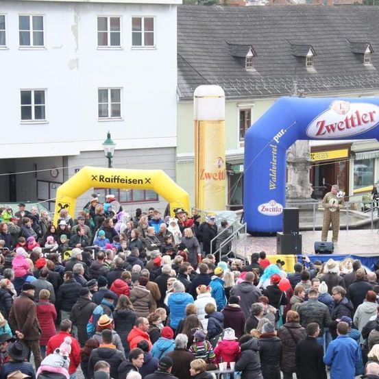 Eine Menschenmenge versammelt sich bei einer Veranstaltung im Freien mit Bühne, Lautsprechern und Bannern. Ein Mann steht auf der Bühne und hält einen Fußball. Im Hintergrund befinden sich Gebäude und ein Straßenlaterne.