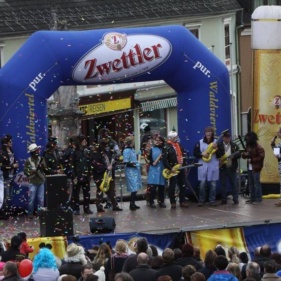 Eine Musikband tritt vor einem Publikum auf der Bühne auf, mit Konfetti und einem blauen Bogen, auf dem Zwettler steht.