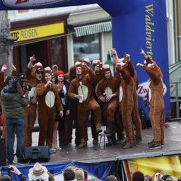 Eine Gruppe von Menschen in Affenkostümen tritt auf der Bühne auf, hebt die Hände und hält Tassen. Dahinter sieht eine Menge zu, und es gibt Banner und einen Baum im Hintergrund.