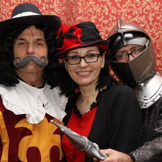 Drei Menschen in mittelalterlichen Kostümen posieren für ein Foto. Ein Mann mit Schnurrbart und eine Frau in einem roten Shirt stehen neben einem Ritter in Rüstung.