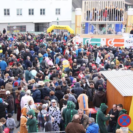 Eine große Menschenmenge versammelt sich im Freien, einige in Kostümen, und einem gelben Bogen mit der Aufschrift 'X Raiffeisen X'. Es gibt eine Holzkonstruktion mit Dach und einem gelben Banner. Menschen stehen auf der Bühne und einige schauen zu.
